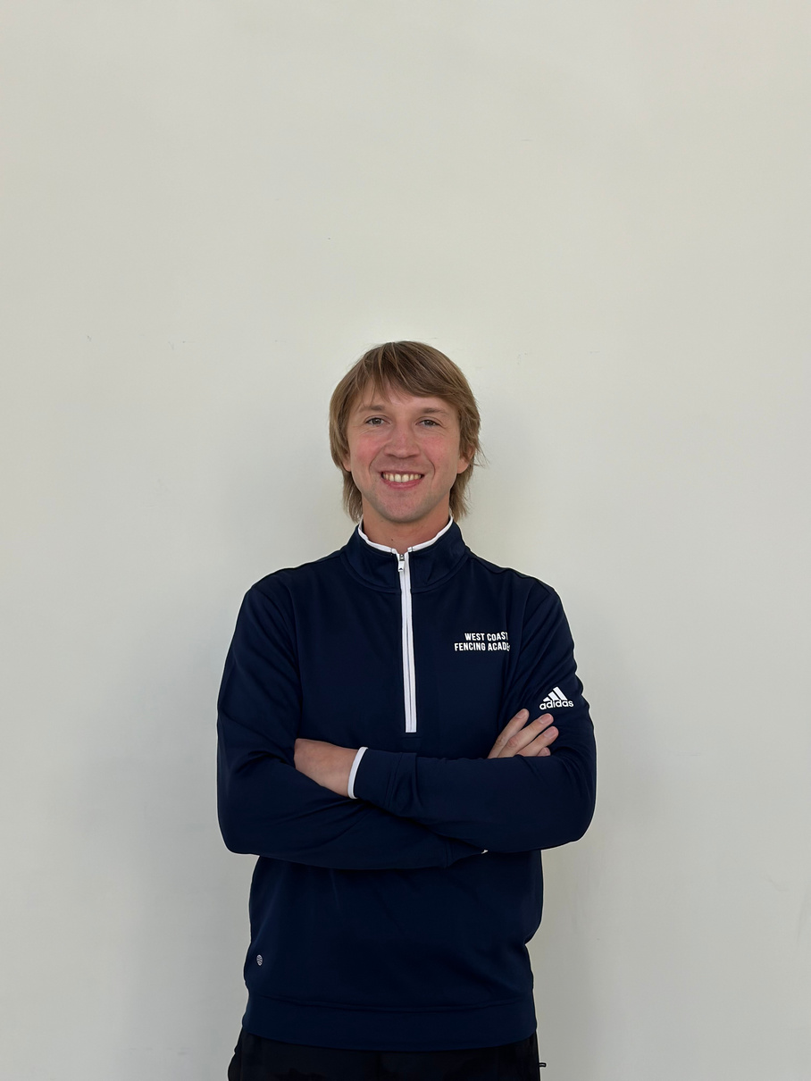 a person with their arms crossed standing in front of a white wall. Coach Nikolay Kovalev posing at West Coast Fencing Academy for Saber fencing