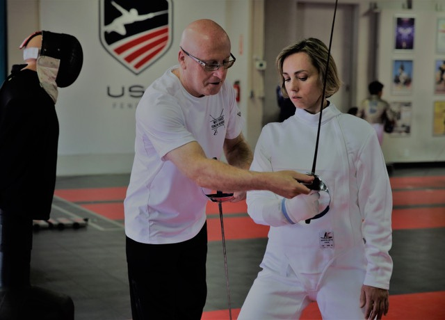 Coach gherman stone teaching a student dressed in saber fencing gear the basics of fencing in en garde stance with a saber in west coast academy gym