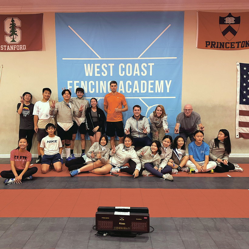 a group of people posing for a photo in front of the west coast fencing academy sign. a group of kids saber fencers wearing saber fencing gear in front of west coast fencing academy flag. 