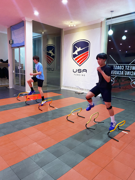 two individuals are doing jumping exercises in a gym. Two teenage saber fencer boys doing agility and plyometric exercises at the fencing gym