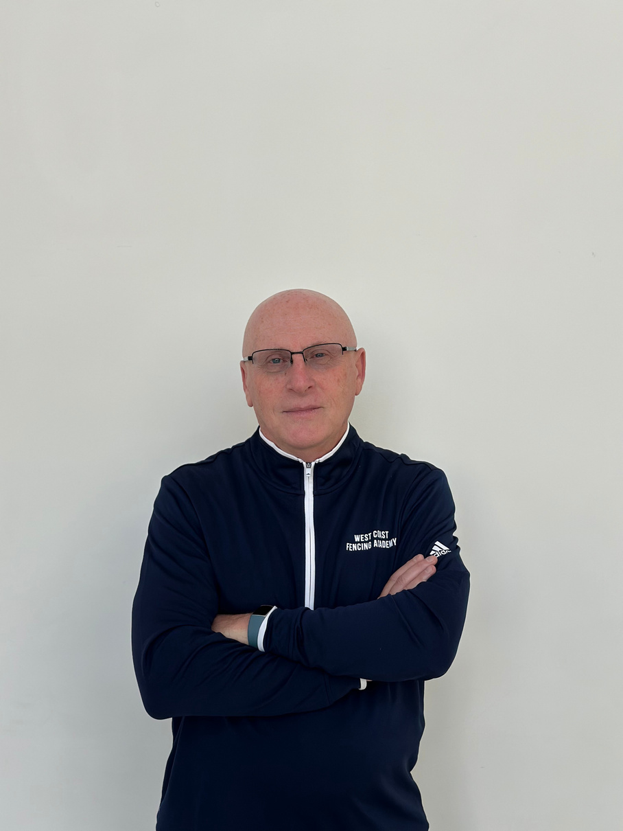 a person with glasses standing in front of a white wall.  Coach Gherman Stone posing at West Coast Fencing Academy